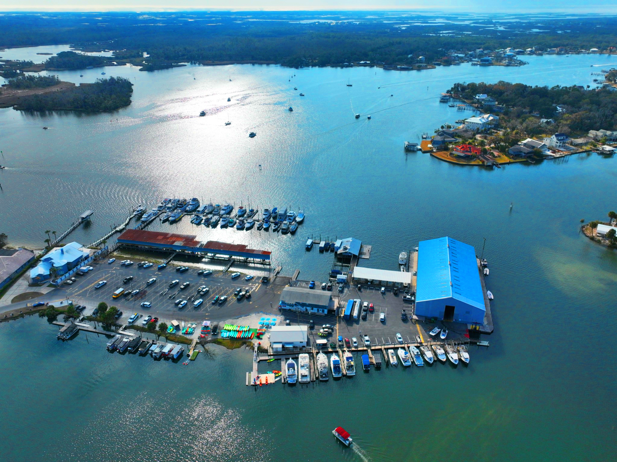 Aerial view of marina with boats, parked cars, and buildings near a large body of water.